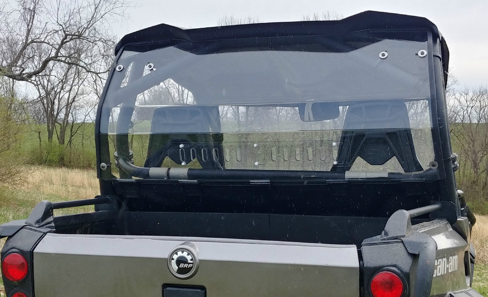 A close-up rear view of a gray Can-Am featuring the Commander/Max Lexan Rear Windshield (2011-2020) reveals its cargo bed. Trees, dry grass, and a cloudy sky create an outdoor rural backdrop.