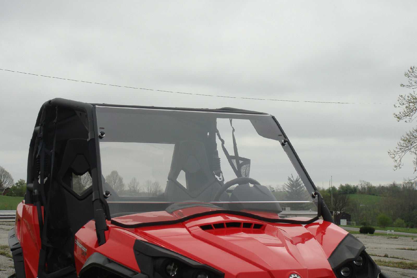 A Can-Am Commander/Max Split Full Lexan Windshield (2011+) is installed on a red off-road utility vehicle parked outdoors with trees, grassy fields, and cloudy skies visible in the background. The vehicle appears empty, showcasing the windshield.