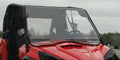 A close-up of a red Can-Am Commander with a 2 Pc General-Purpose Windshield (vent and clamp options) and black roof, parked outdoors on a cloudy day. The driver’s seat, steering wheel, grass, and trees are visible in the background.