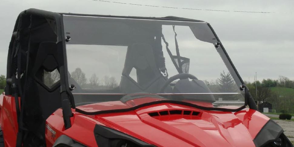 A close-up of a red Can-Am Commander with a 2 Pc General-Purpose Windshield (vent and clamp options) and black roof, parked outdoors on a cloudy day. The driver’s seat, steering wheel, grass, and trees are visible in the background.