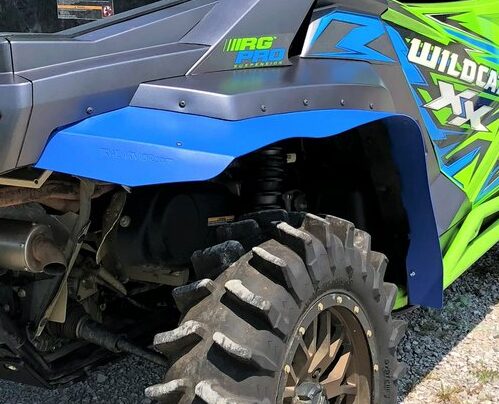 Close-up of Trail Armor Textron Wildcat XX Mud Flap Fender Extensions installed on a rugged off-road vehicle, featuring thick knobby tires on gravel and bright blue/green graphics with WILDCAT XX and RG PRO visible on the side panel.