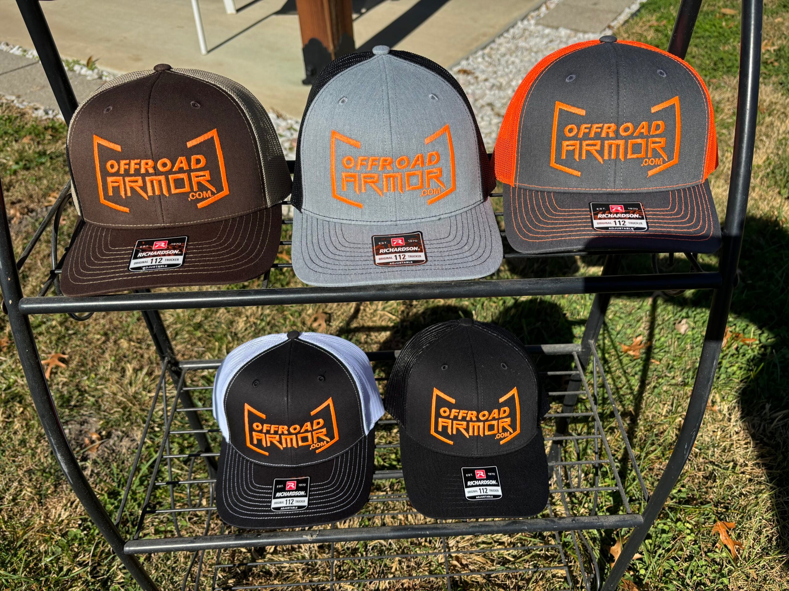 Five Offroad Armor Richardson Snapbacks in brown, gray, black, and mixed designs are arranged on a metal rack outdoors, displayed on two shelves in sunlight. Each hat features an orange embroidered logo on the front panel. Grass is visible below.