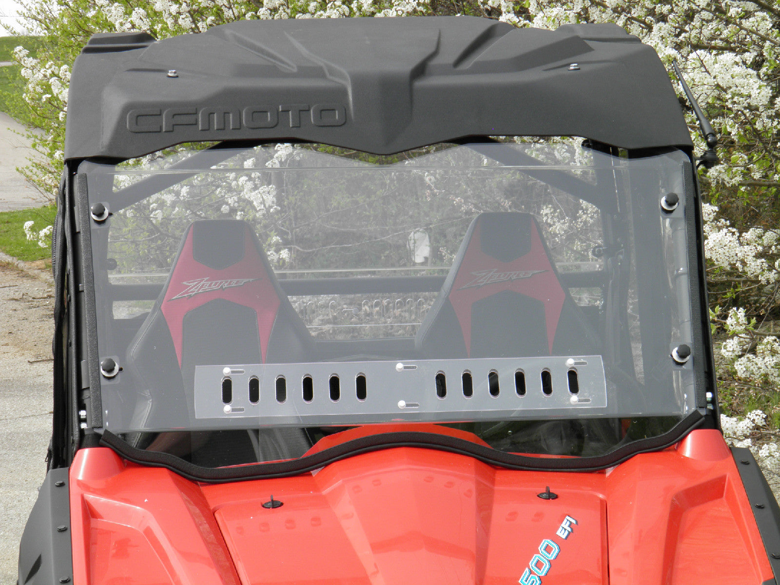 A close-up shows a Full Lexan Windshield on a red CF Moto Z Force 500 with two black and red seats and a black roof. White-blossomed trees are in the background, with outdoor scenery reflected on the windshield.
