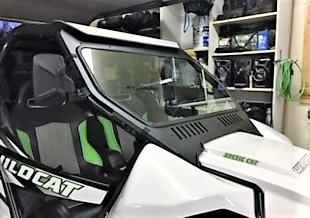 A white Arctic Cat Wildcat with green and black seats and a bold “WILDCAT” logo is parked indoors near shelves of gear. It features an Arctic Cat Wildcat Vented Windshield With D.O.T Stamp.