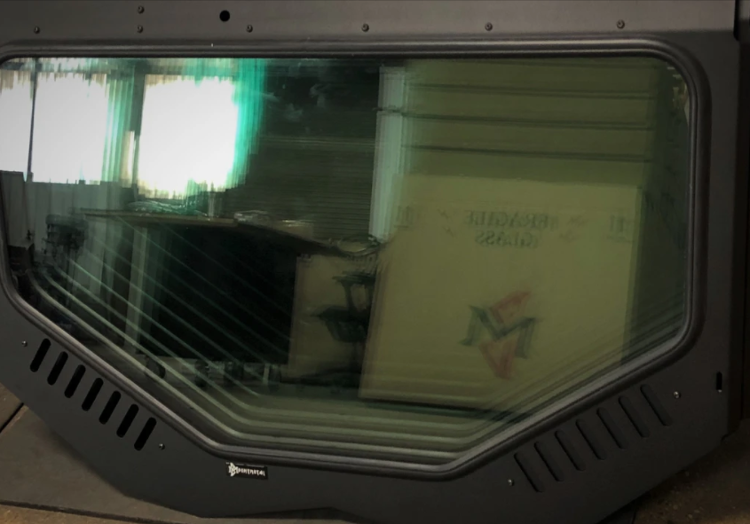 A large, tinted Replacement Glass with a D.O.T stamp leans against a wall, reflecting bright light and stacked cardboard boxes. The black frame features ventilation slits and a small Dynomax sticker.