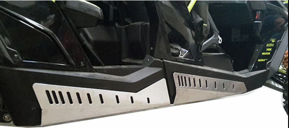 Close-up of the lower side panels on a Can Am Maverick 1000 4 Seater, featuring Rock Slider guards in silver metal with vertical slits and angular styling. Some controls and labels are visible above the sliders.