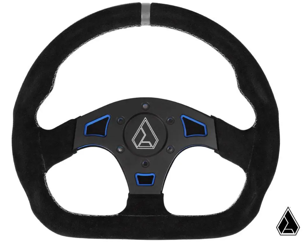 The Suede Ballistic D UTV Steering Wheel features a black suede finish, blue accents, a metallic geometric-logo hub, three spokes, white edge stitching, and a small matching logo in the bottom right corner.