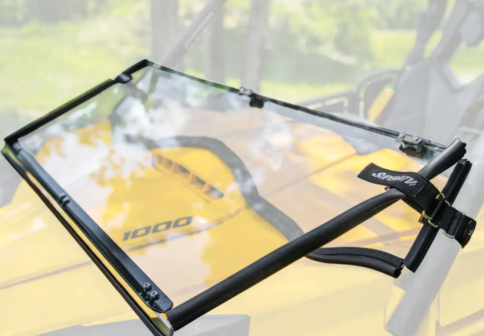 A Can-Am Commander Scratch Resistant Flip Down Windshield with a black metal frame appears close-up against a blurred yellow off-road vehicle marked 1000. A SuperATV-branded mounting strap attaches to the top right corner of the frame.