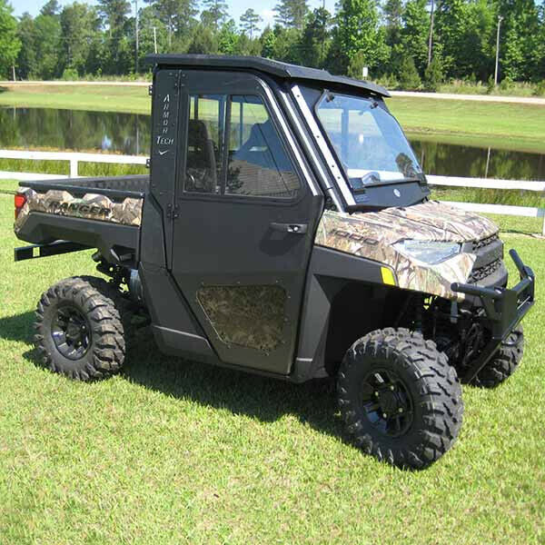 A Polaris Ranger XP 1000 with Armor Tech Full Steel Doors featuring sliding windows is parked on grass near a pond and white fence. It has camouflage door and hood accents, large off-road tires, tinted windows, and a protective roof.
